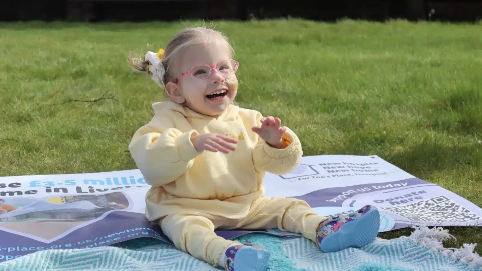 Photo showing a smiling child sitting on a mat and Zoe's Place banner in the grass.