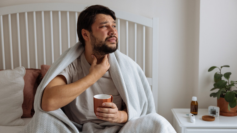 A man sitting up in bed holding his sore throat with one hand and a mug in another