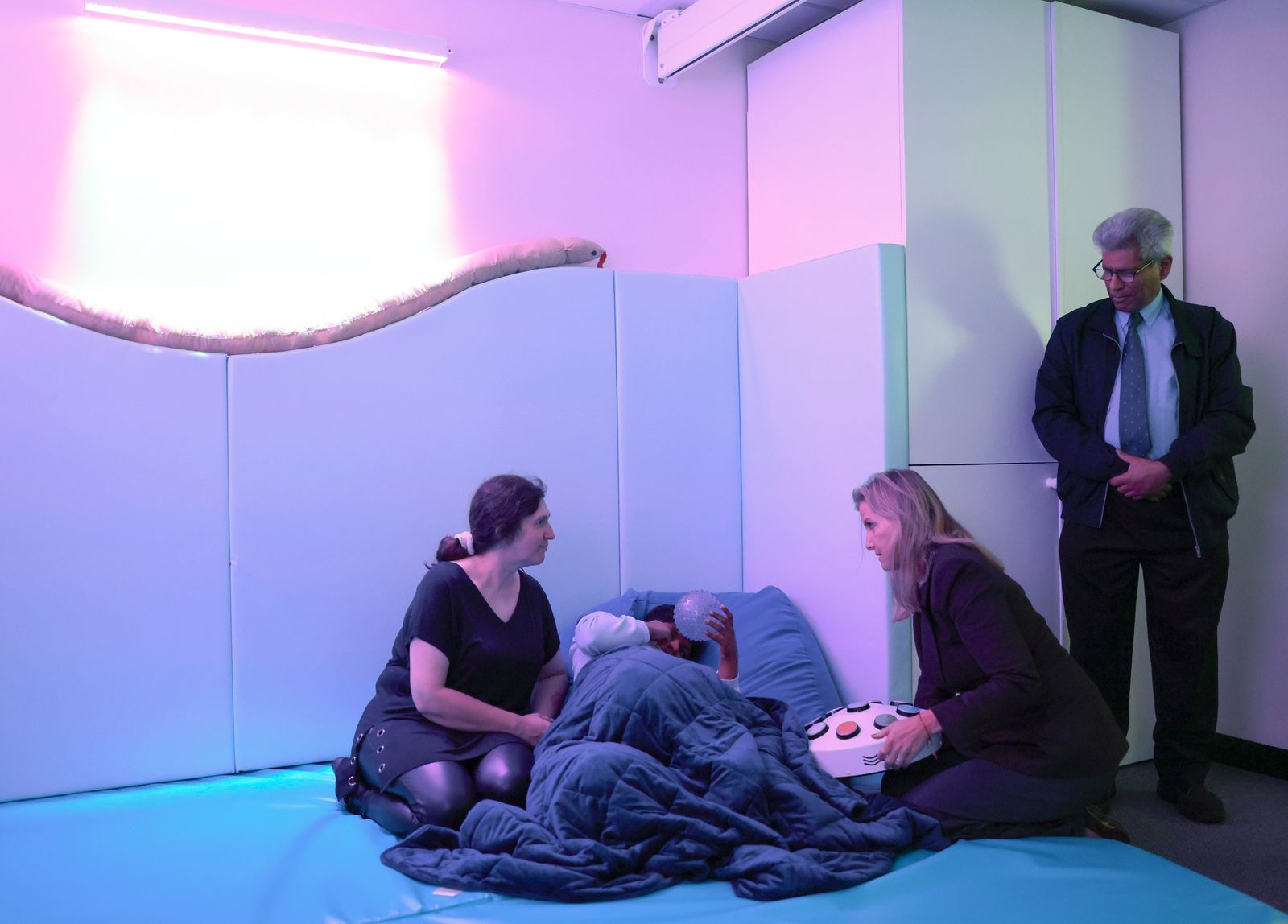 sensory room with blue floor soft gray forms along wall with curving top snake atop curve purple lighting child relaxes...