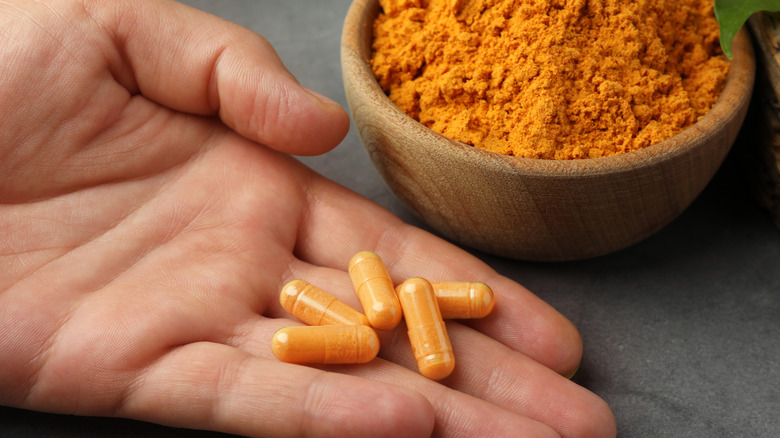 A woman's hand holding turmeric supplement pills next to a bowl of turmeric powder